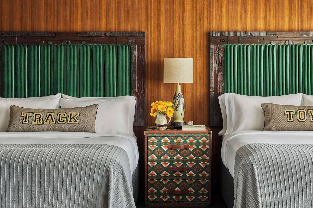 A hotel room with two beds, each with a pillow featuring the word "TRACK" and "TOWN" respectively. A nightstand with a lamp and a vase of flowers is between the beds.