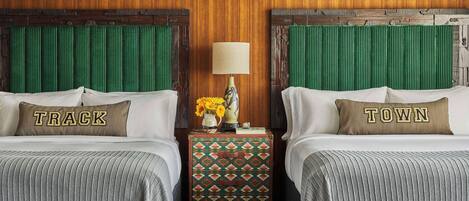 A hotel room with two beds, each with a pillow featuring the word "TRACK" and "TOWN" respectively. A nightstand with a lamp and a vase of flowers is between the beds.