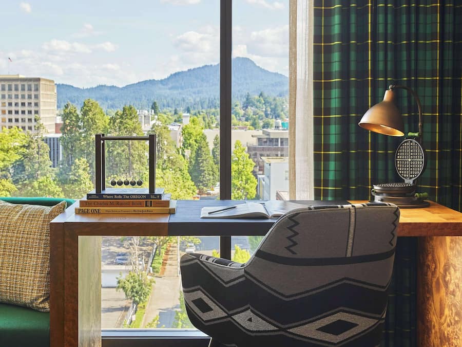 Habitación con escritorio, una silla, una lámpara y vistas a montañas y edificios.