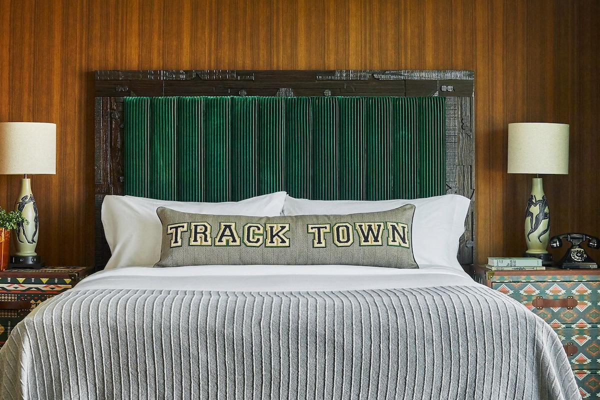 A bed with a green headboard, two bedside lamps, a pillow with 'TRACK TOWN' text, and patterned side tables.