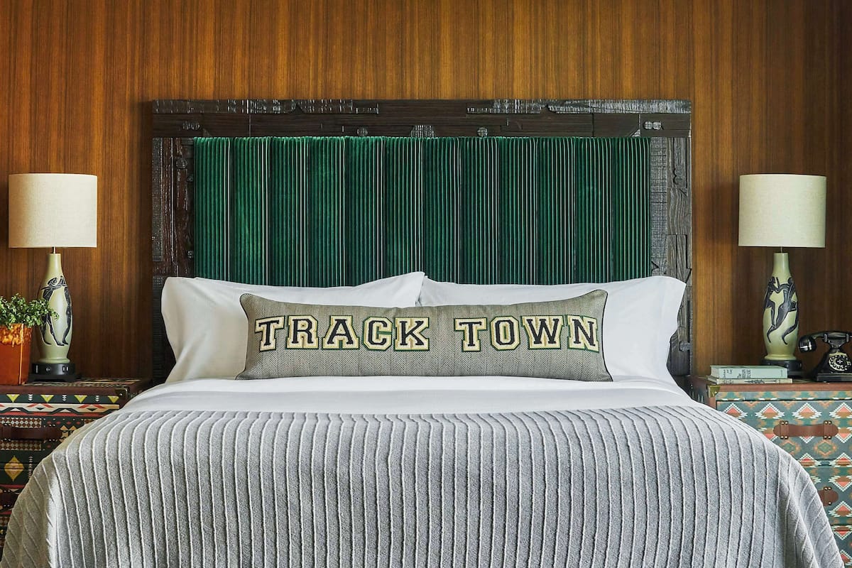 A bed with a green headboard, two bedside lamps, a pillow with "TRACK TOWN" text, and patterned bedding.
