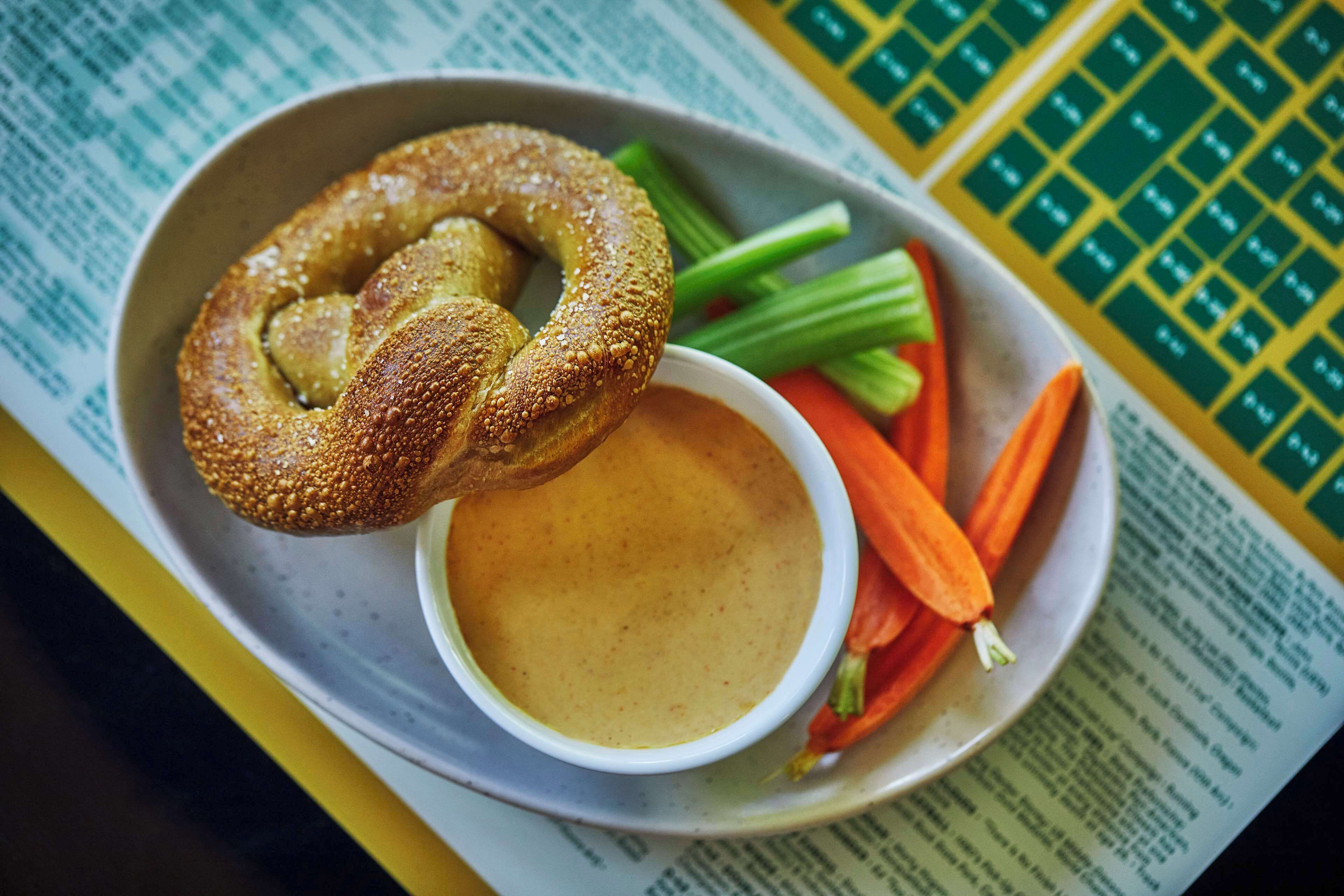 Prato com pretzel, aipo, cenouras e uma tigela de molho sobre uma mesa com um menu.