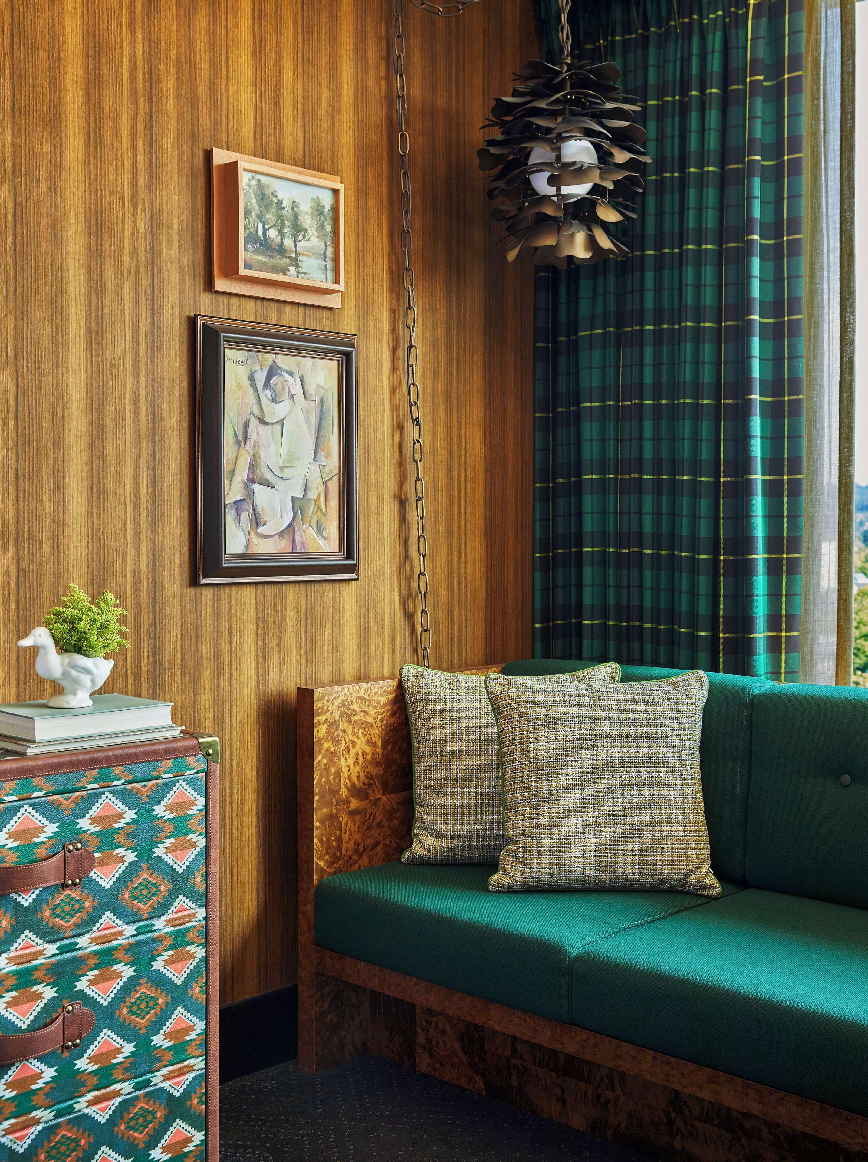 Una habitación acogedora con pared de madera, un sofá verde, cojines estampados, una mesita con una planta y un libro, y una lámpara colgante.