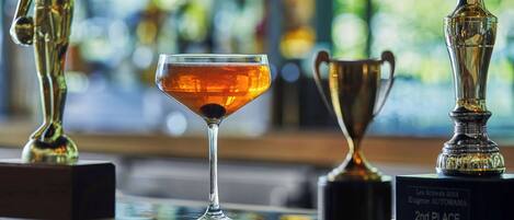 Un vaso de bebida color ámbar sobre una superficie reflectante, con trofeos y una estatuilla al fondo.