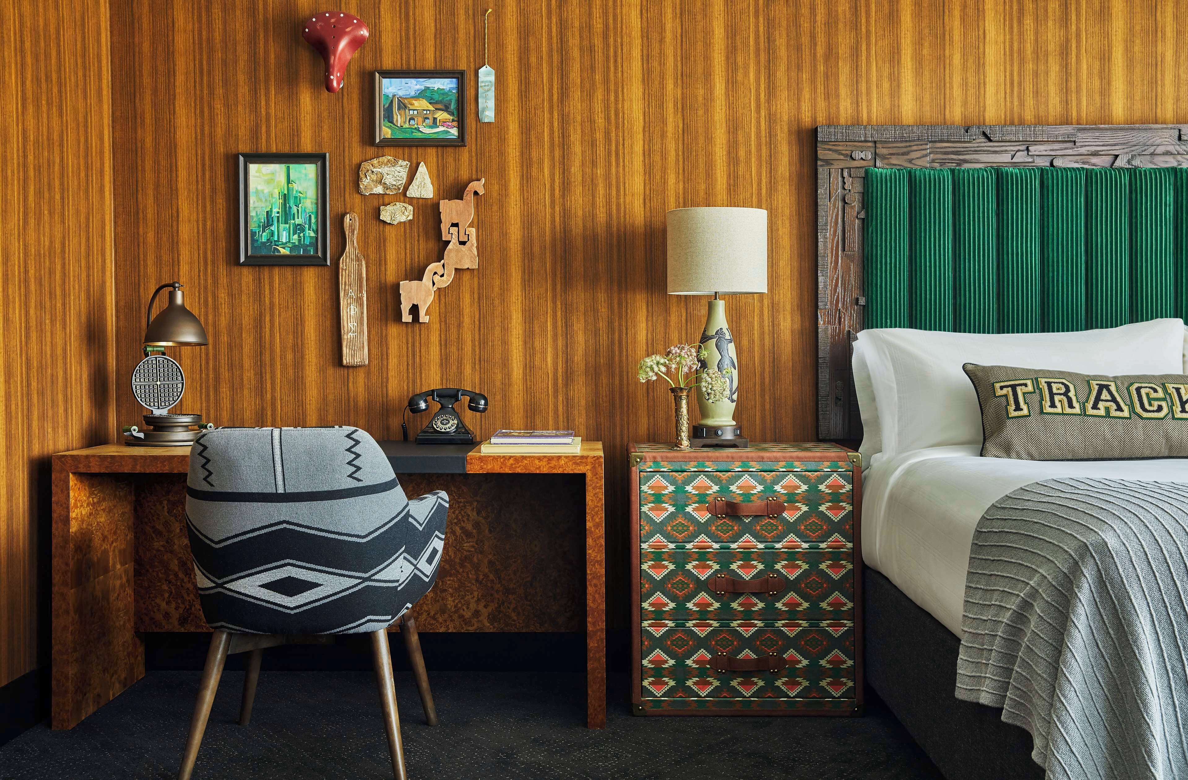 A wooden-paneled room with a bed, a desk, a chair, and a nightstand with a lamp.