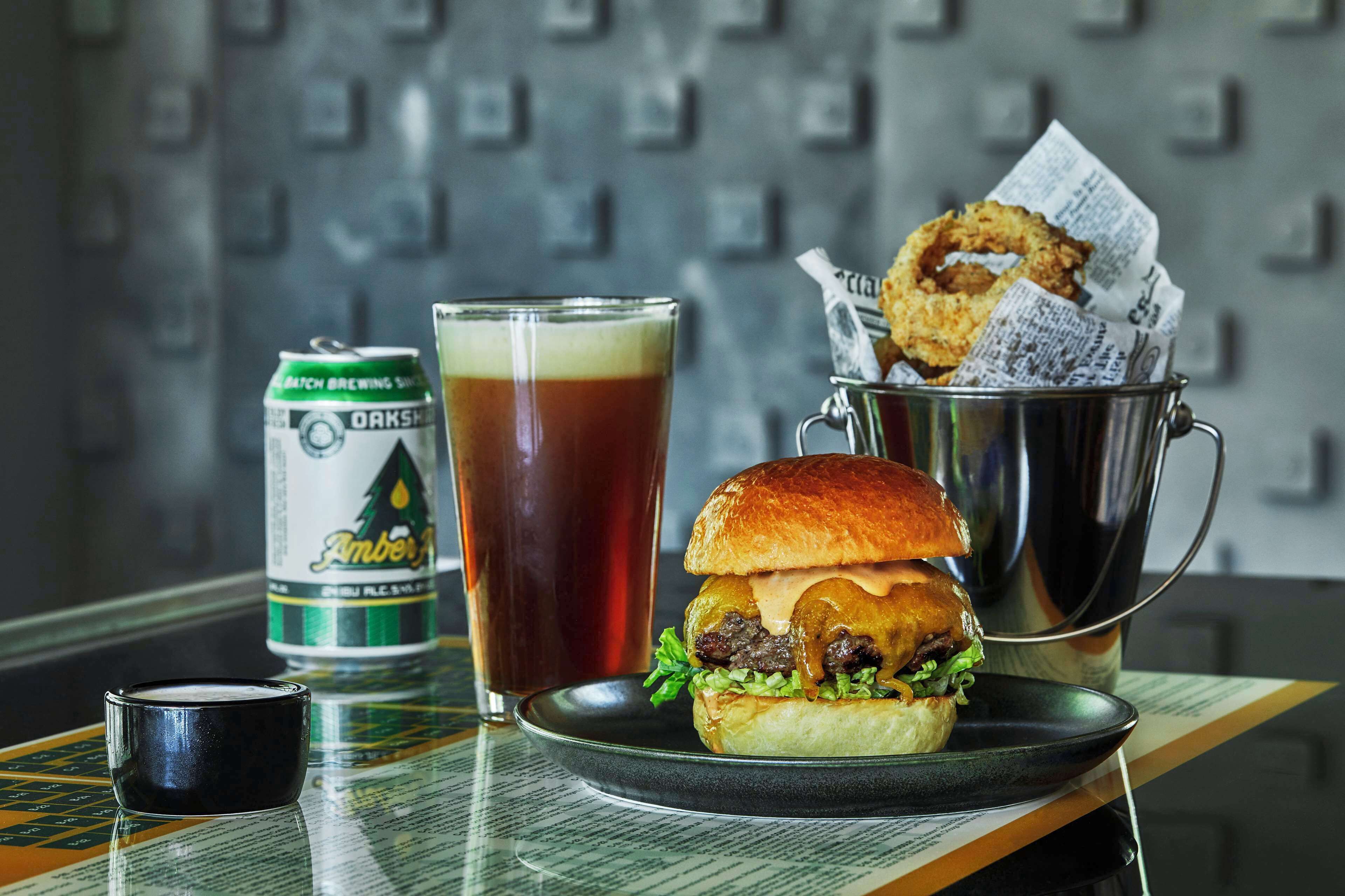 A burger, a glass of beer, and a can of beer on a table with a newspaper and a small cup.