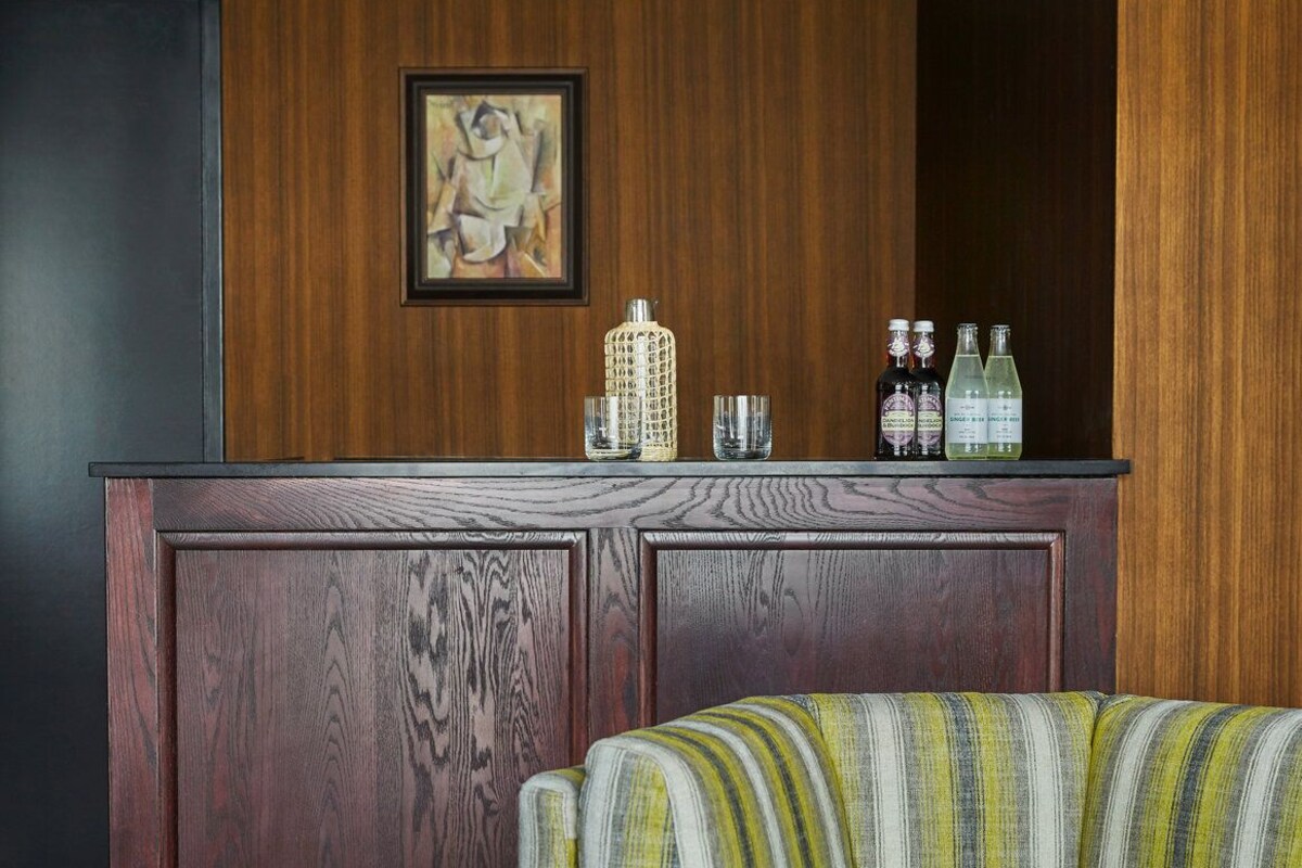 A room with a wooden panel wall, a framed picture, a bar area with bottles, glasses, and a woven container.