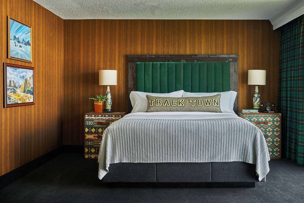 A hotel room with a wooden paneled wall, a bed with a green headboard, two bedside lamps, and a window with a view of greenery.