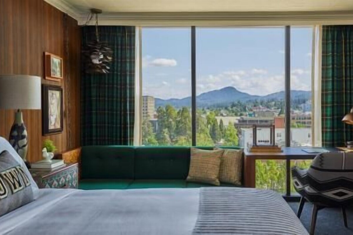 A hotel room with a large window, a bed, a desk, and a chair.