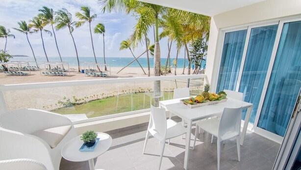 Un balcón con vistas a la playa, muebles blancos y una mesa con fruta.