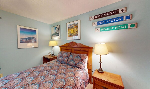 A bedroom with a bed, two bedside tables, a nightstand, and wall-mounted signs reading "DETONATOR," "PROSPECTOR," and "HEADIN' HOME." - Close to Mountain Activities | Fireplace | WiFi | Wren 101 (Angel Fire)