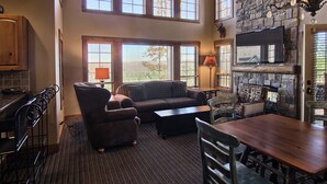 A spacious living area with a large window, a stone fireplace, and a television mounted on the wall. - Jump onto the Disciples 8 Chairlift - 7317 Creekside Condo, Ski in/Ski out (Boyne City)