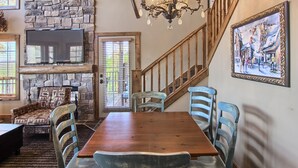 A dining area with a wooden table and blue chairs, a stone fireplace, a television mounted on the wall, and a staircase with wooden railings. - Jump onto the Disciples 8 Chairlift - 7317 Creekside Condo, Ski in/Ski out (Boyne City)