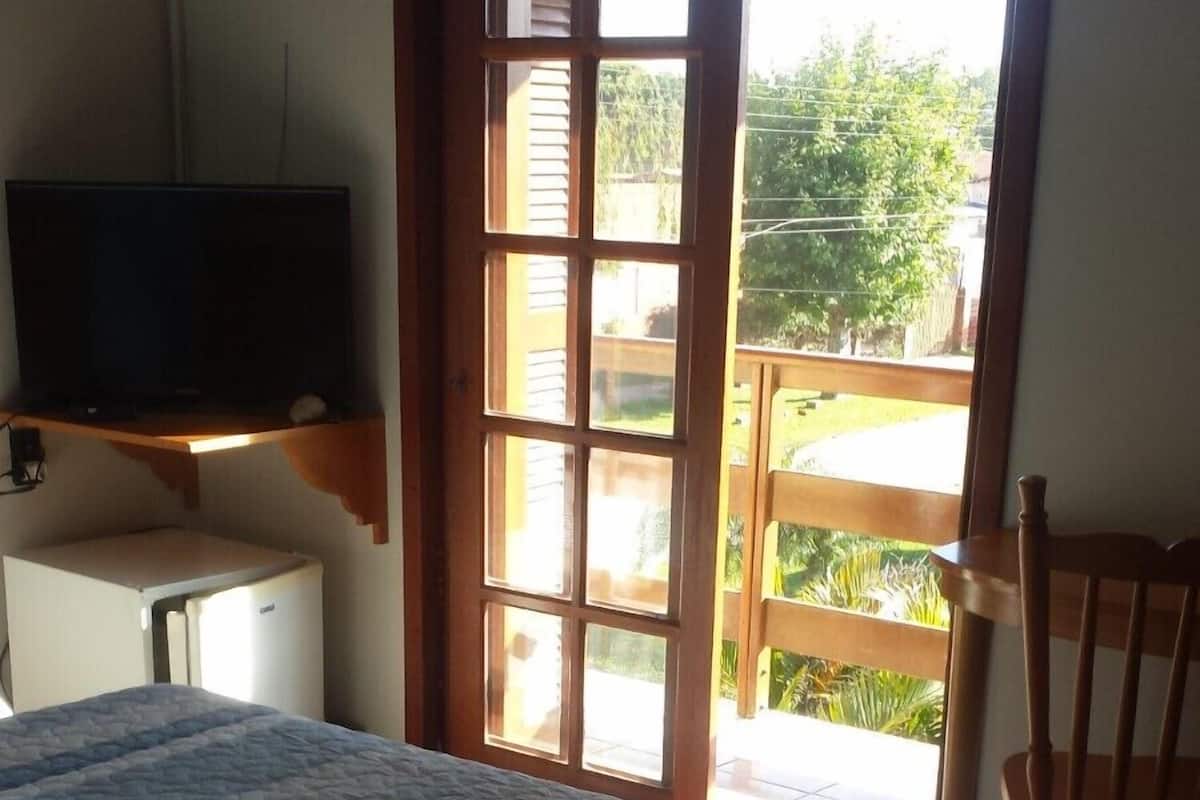 Un dormitorio con una cama, un televisor en un estante, una silla de madera y una puerta corrediza de vidrio que da a un espacio al aire libre.