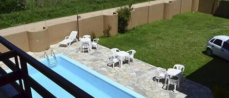 A swimming pool with lounge chairs and a stone deck, surrounded by a low wall and a grassy area.