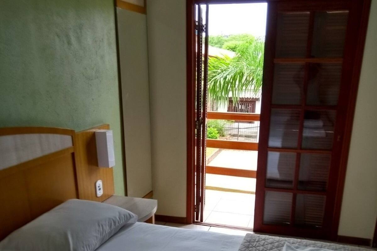 Habitación de hotel con cama, mesita de noche, aire acondicionado instalado en la pared y puerta corrediza de vidrio que da a un espacio exterior con vegetación.