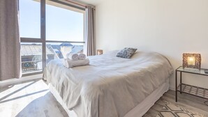 A hotel room with a large bed, a bedside table with a lamp, a view of the city through a large window, and a balcony with a railing. - Apt Centro Bariloche Cochera Incluida II (San Carlos de Bariloche)