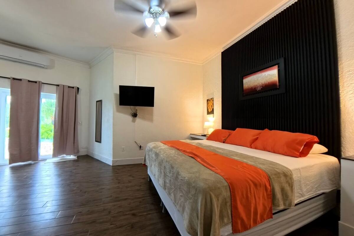 Egy hálószoba, amelyben van egy ágy, egy mennyezeti ventilátor, egy televízió és egy ablak, amelyen függöny van.