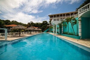 A swimming pool with a clear blue water, surrounded by a white fence, and a building with a balcony in the background. - West End Dive Resort (Roatan)
