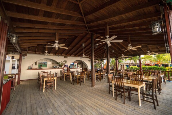 Un área de comedor al aire libre con mesas y sillas de madera, ventiladores de techo y una barra.
