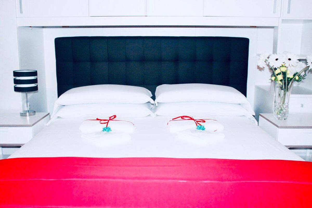 A neatly made bed with white linens and a red blanket, a dark headboard, and a bedside table with a lamp and a vase of flowers.