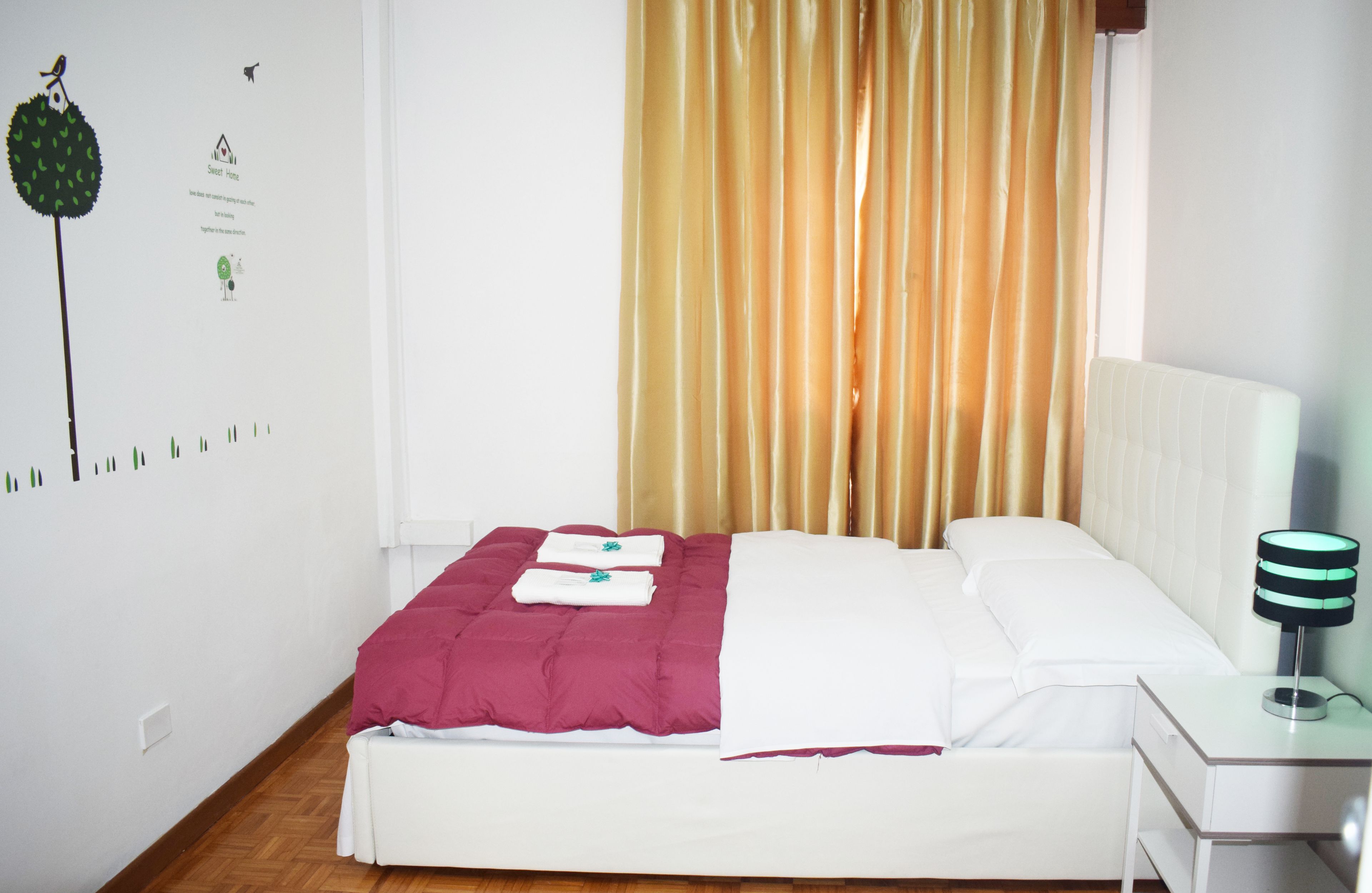 A neatly made bed with a white and maroon bedspread, a white pillow, and a folded towel. There is a bedside table with a lamp, and a window with curtains.