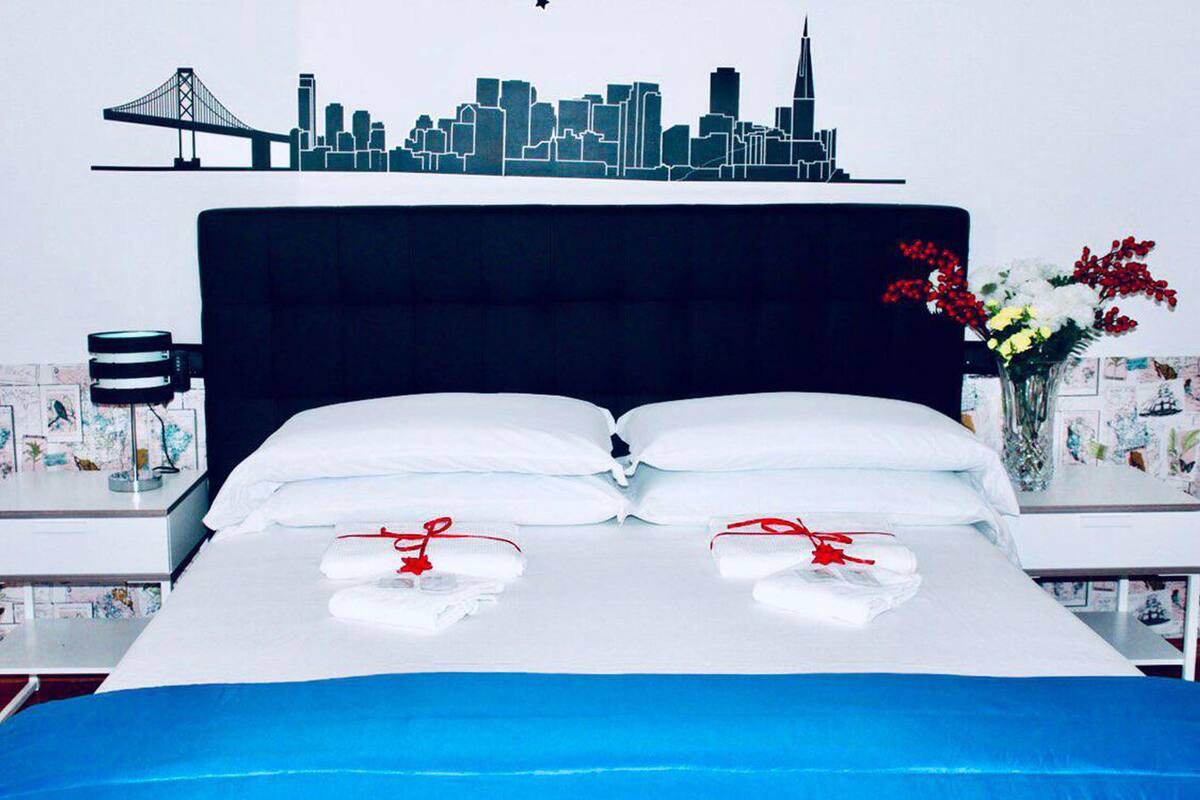 A hotel room with a bed, a city skyline mural, a vase with flowers, and a desk with a lamp.