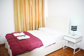 Ein ordentlich bezogenes Bett mit weißem Kopfteil und roter Decke, ein weißer Nachttisch mit Lampe sowie ein Holzboden.