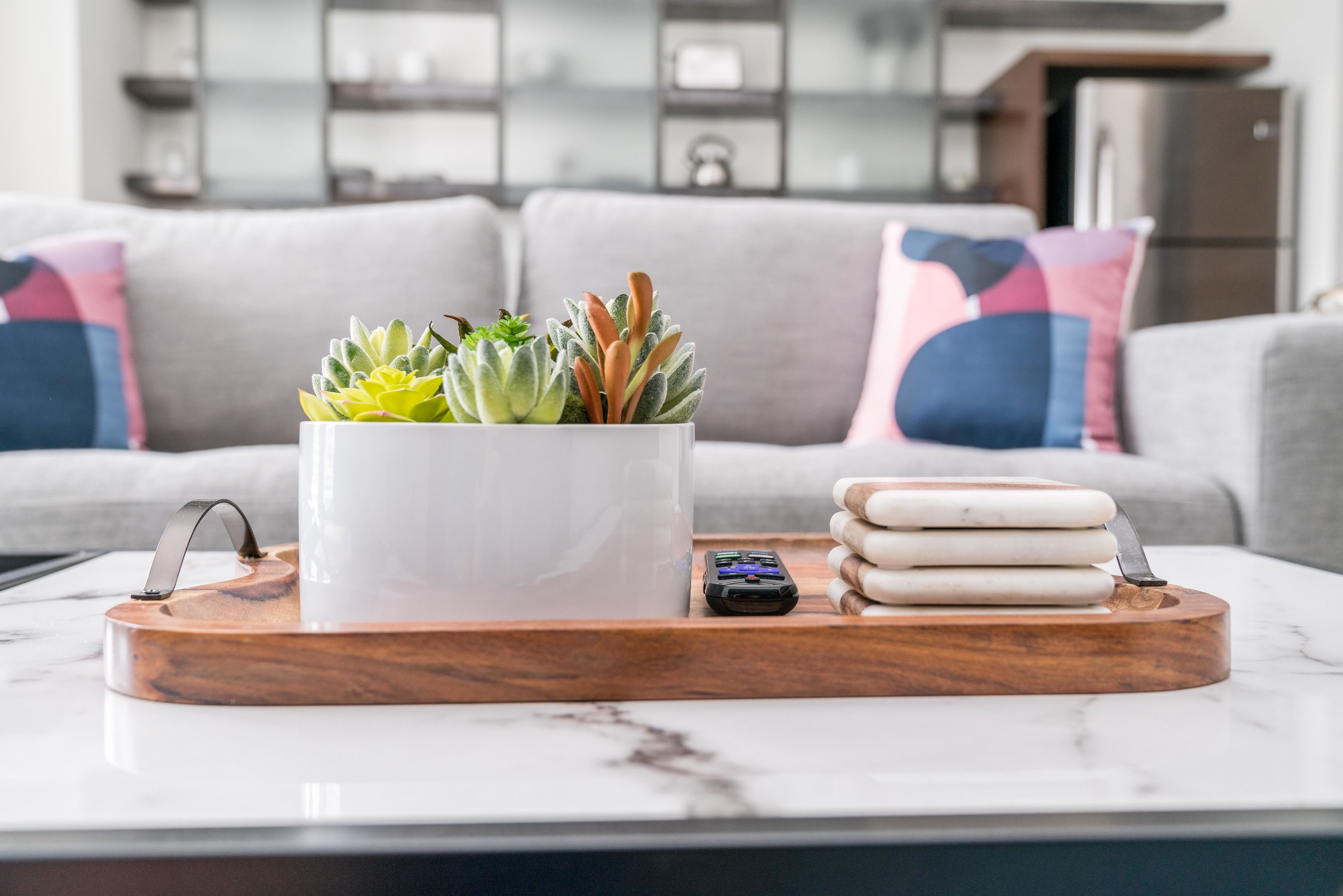 Een moderne woonkamer met een marmeren salontafel, een witte vaas met vetplanten, een klein elektronisch apparaat en een houten dienblad.