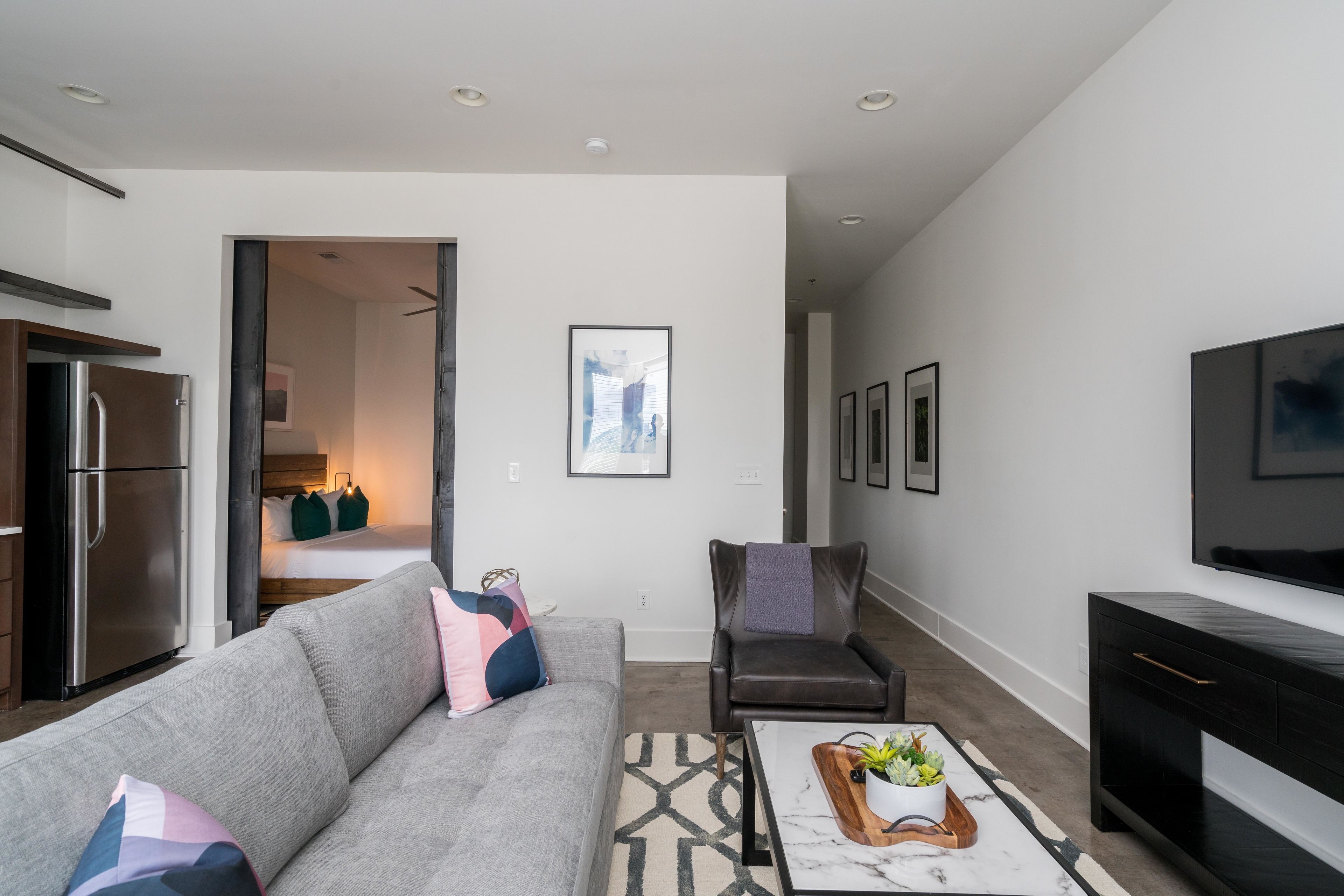 A modern living room with a grey sofa, a coffee table with a tray, a black leather chair, and a TV on the wall.