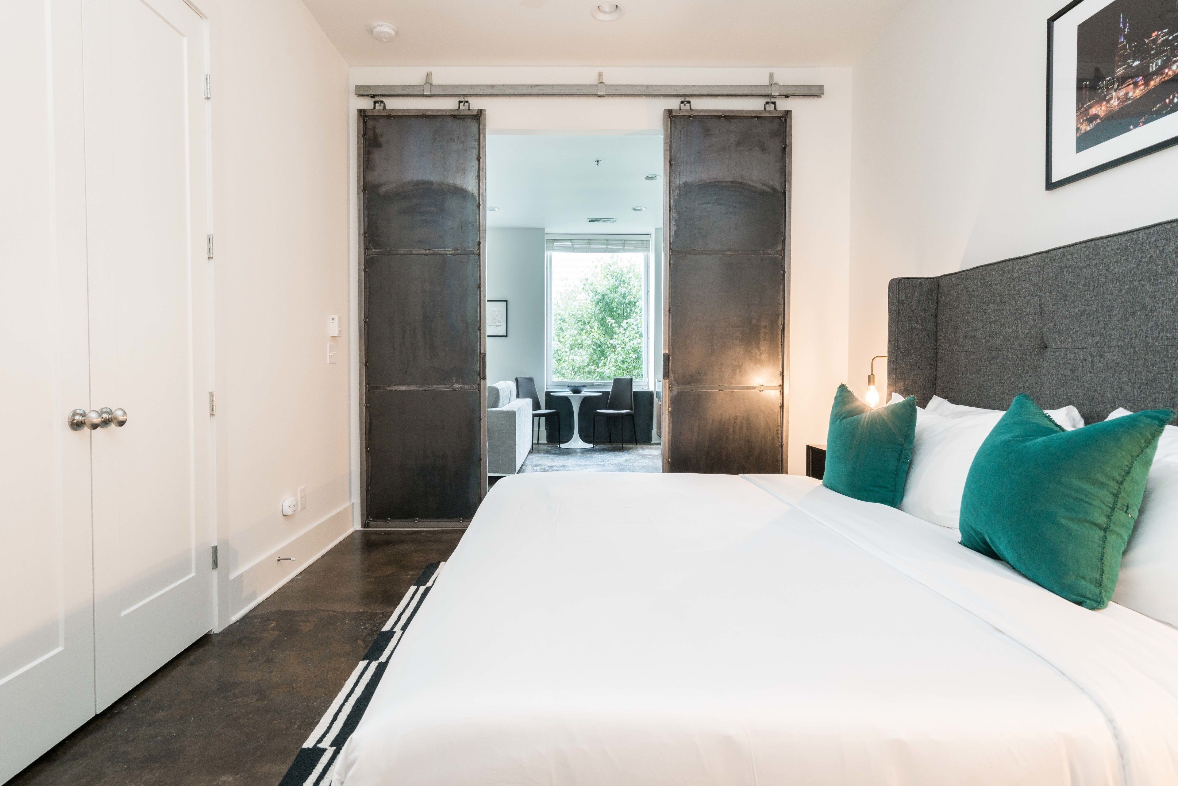 A modern hotel room with a large bed, a view of the outdoors, and a dining area visible through a sliding door.
