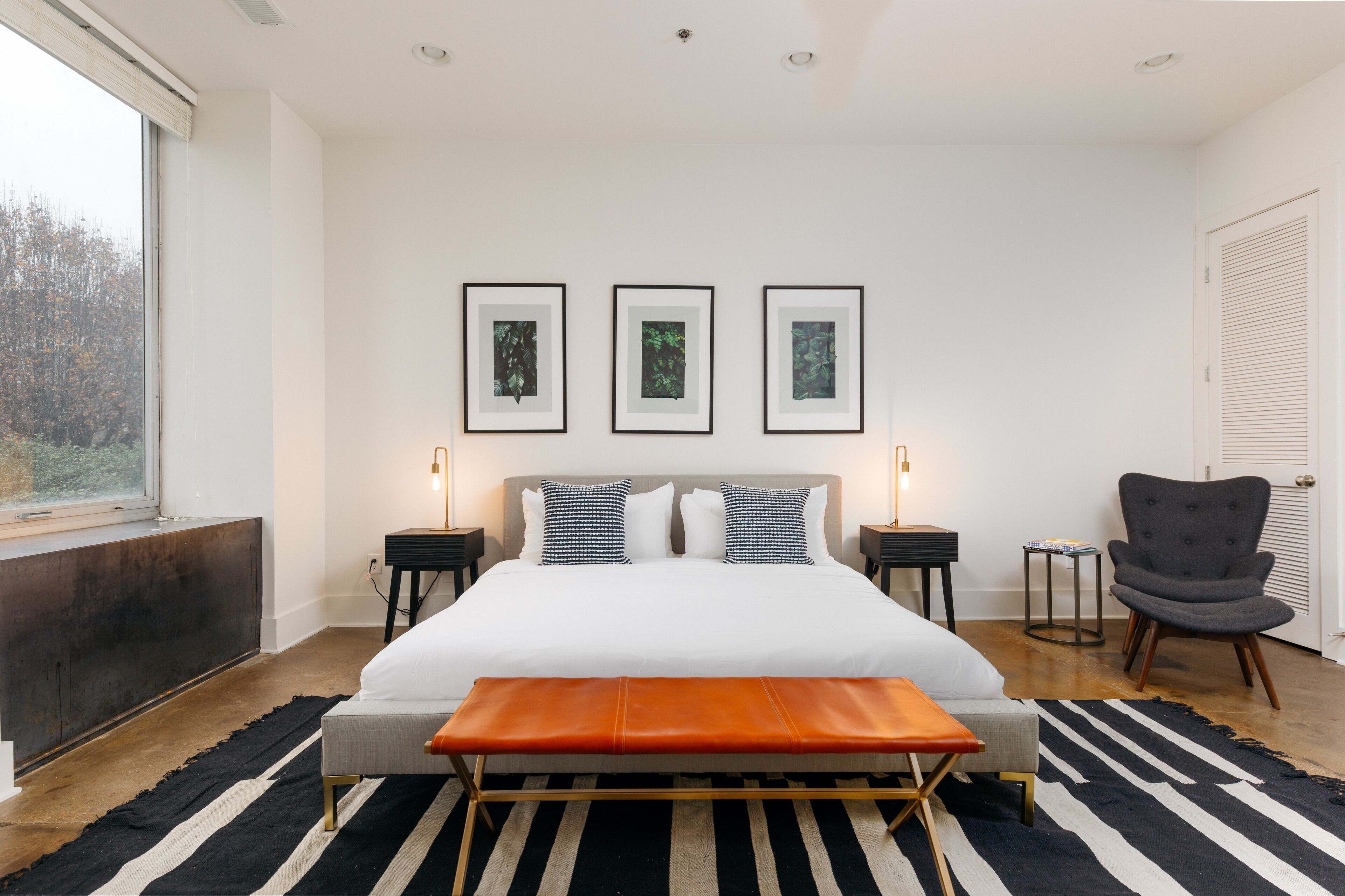A modern bedroom with a large bed, bedside tables, a bench, a chair, and framed artwork on the wall.