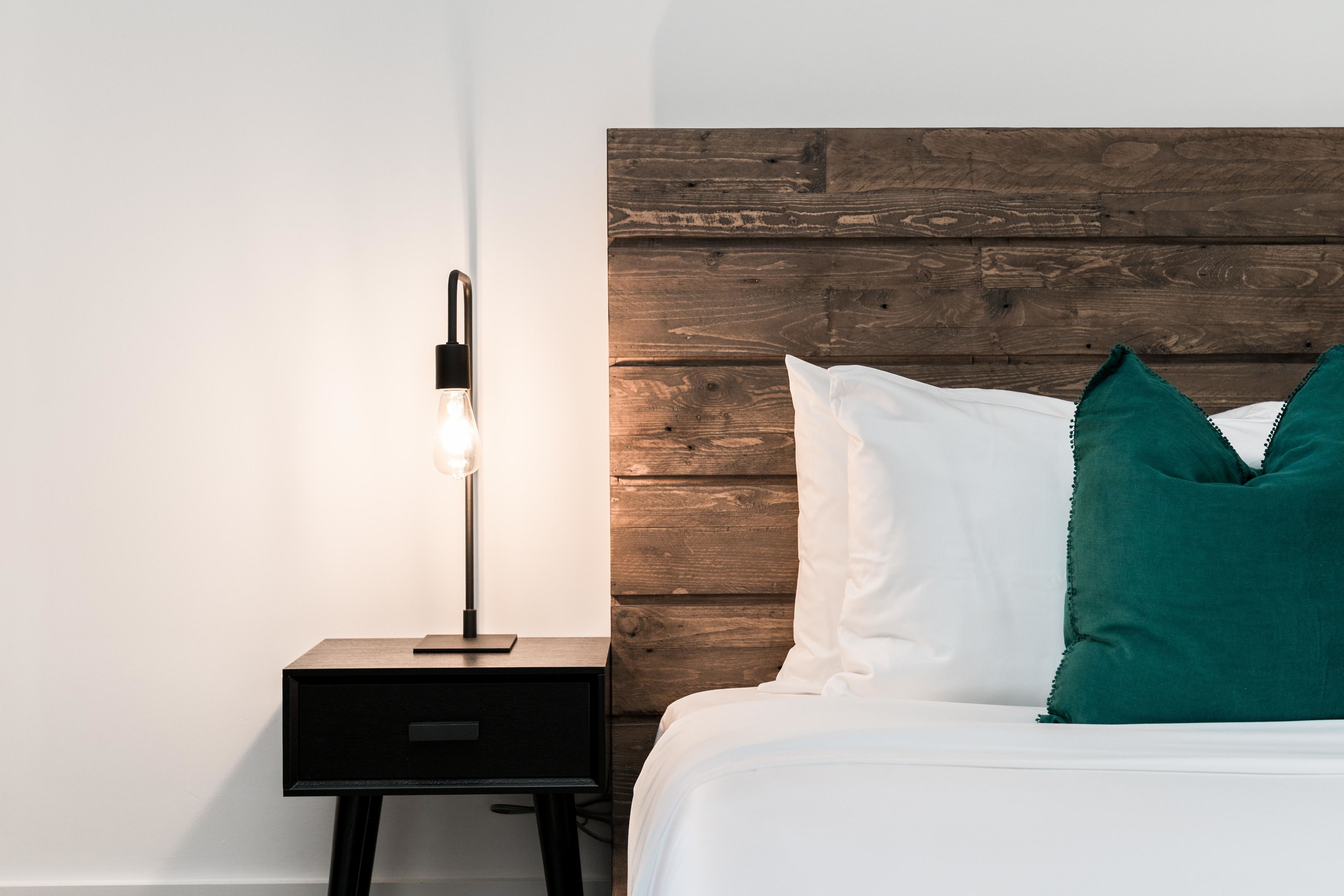 Ein ordentlich bezogenes Bett mit weißen und türkisfarbenen Kissen, einem Holzkopfbrett und einem kleinen Nachttisch mit Lampe.