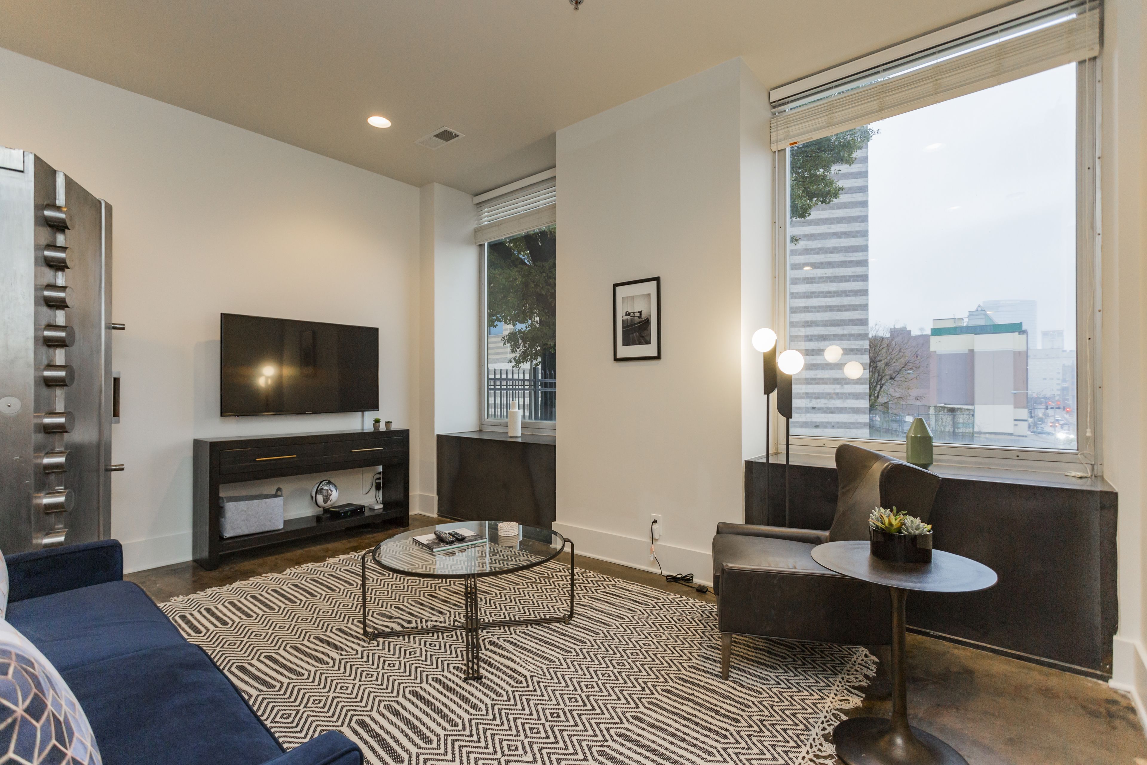 A modern living room with a large TV, a sofa, a coffee table, and a view of the city through a large window.