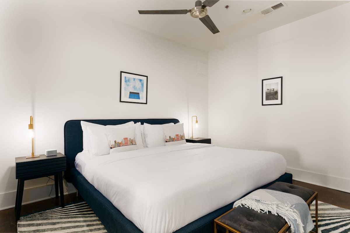 Una cama bien hecha con ropa blanca, cabecera oscura y un pequeño banco al pie. Hay dos mesitas de noche con lámparas, y la habitación tiene un ventilador de techo.