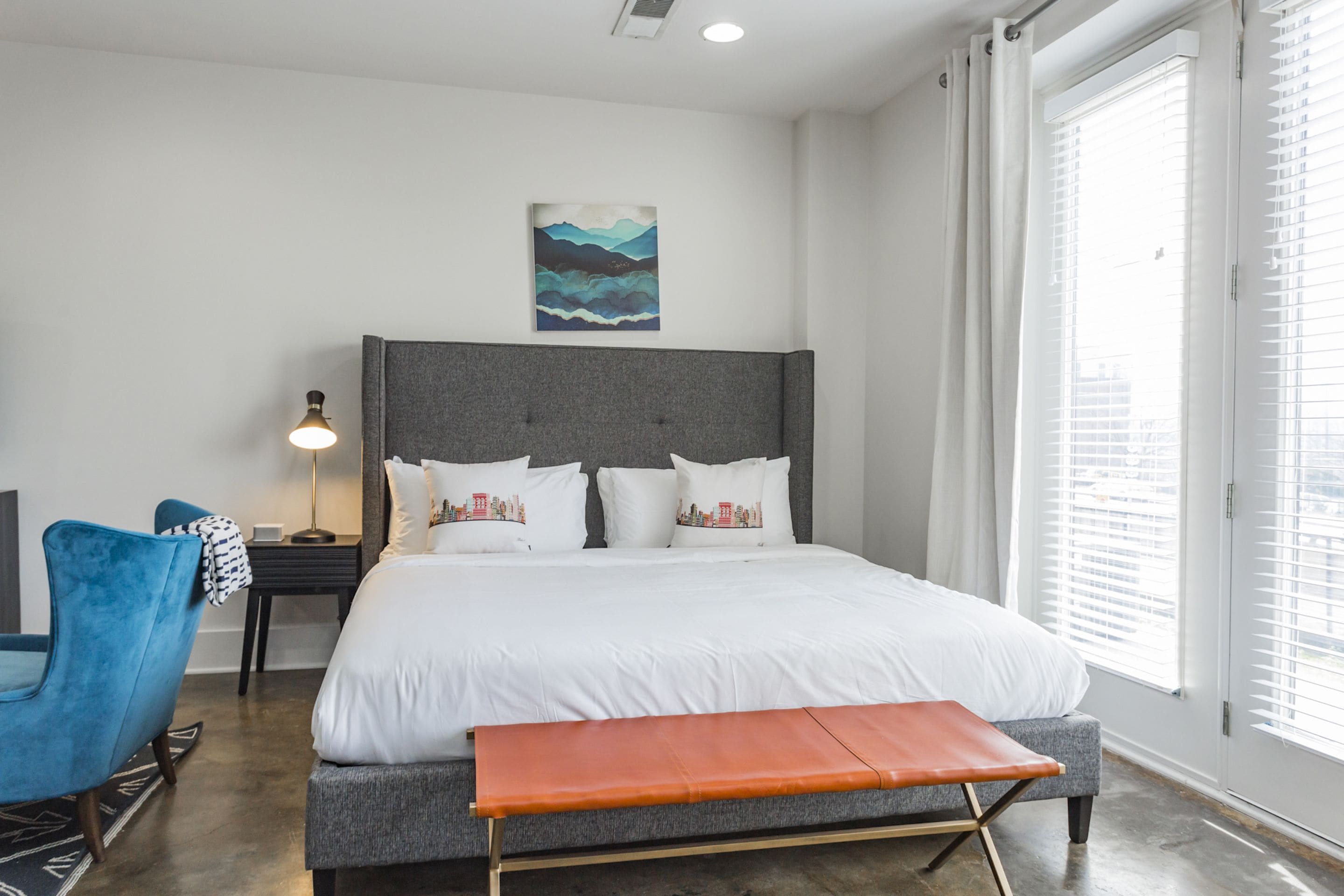 Un dormitorio moderno con una cama grande, cabecera gris, ropa de cama blanca, una silla azul, una mesita de noche con lámpara y una ventana con persianas.