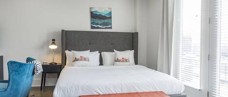 A modern bedroom with a large bed, a grey headboard, white bedding, a blue chair, a bedside table with a lamp, and a window with blinds.
