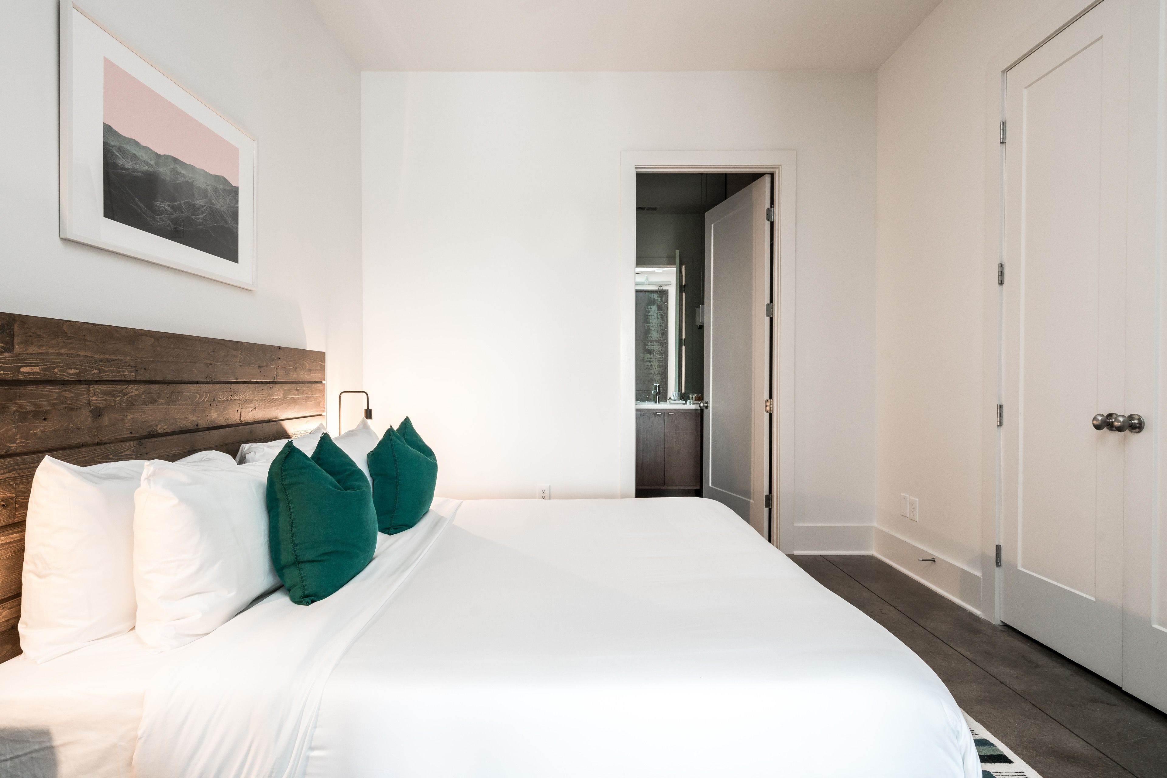 A neatly made bed with white linens and green pillows, a wooden headboard, and a framed picture on the wall.