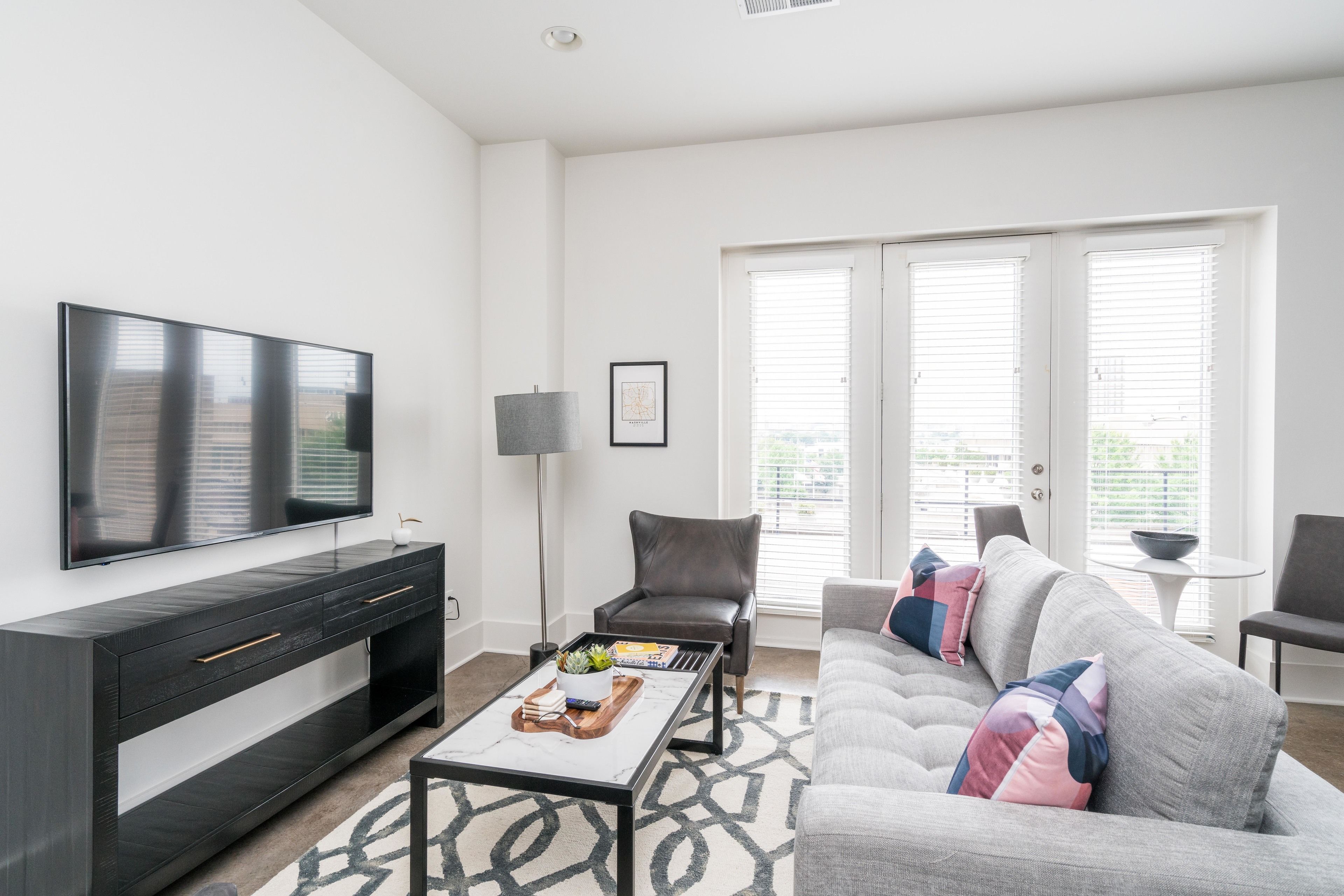 A modern living room with a flat-screen TV, a dark wooden console table, a grey sofa with colorful cushions, a patterned rug, and a floor lamp.