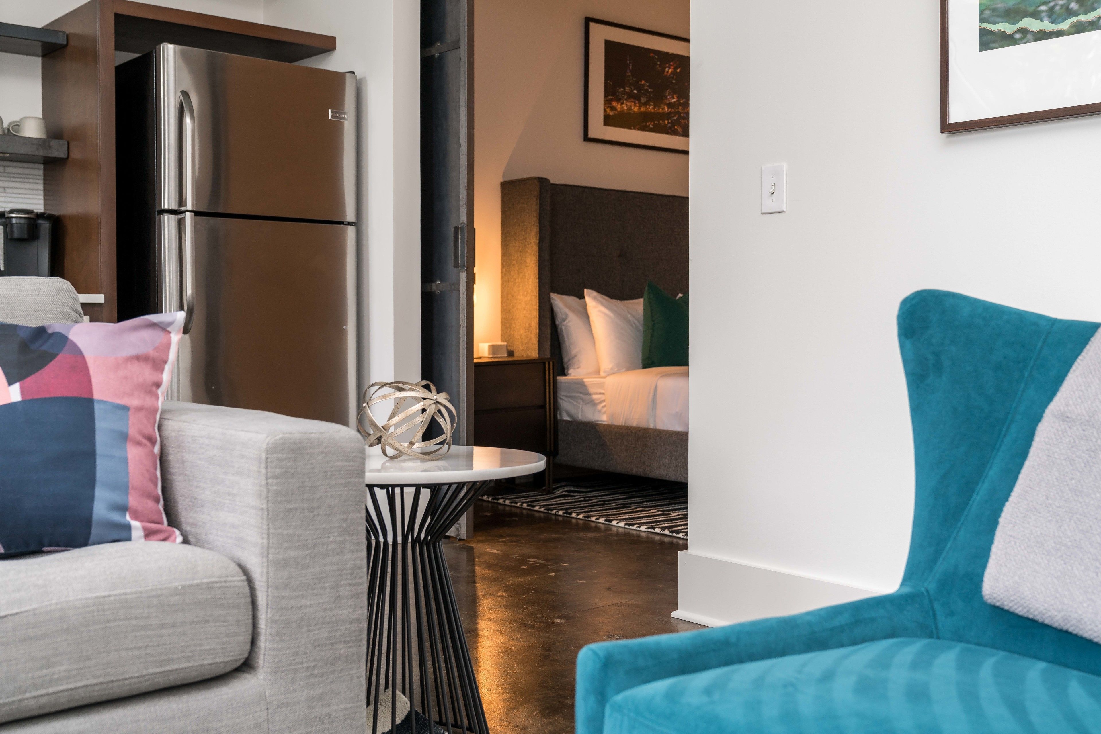 A modern hotel room with a kitchenette, a bed with white bedding, a blue armchair, and a small round table with a decorative item.