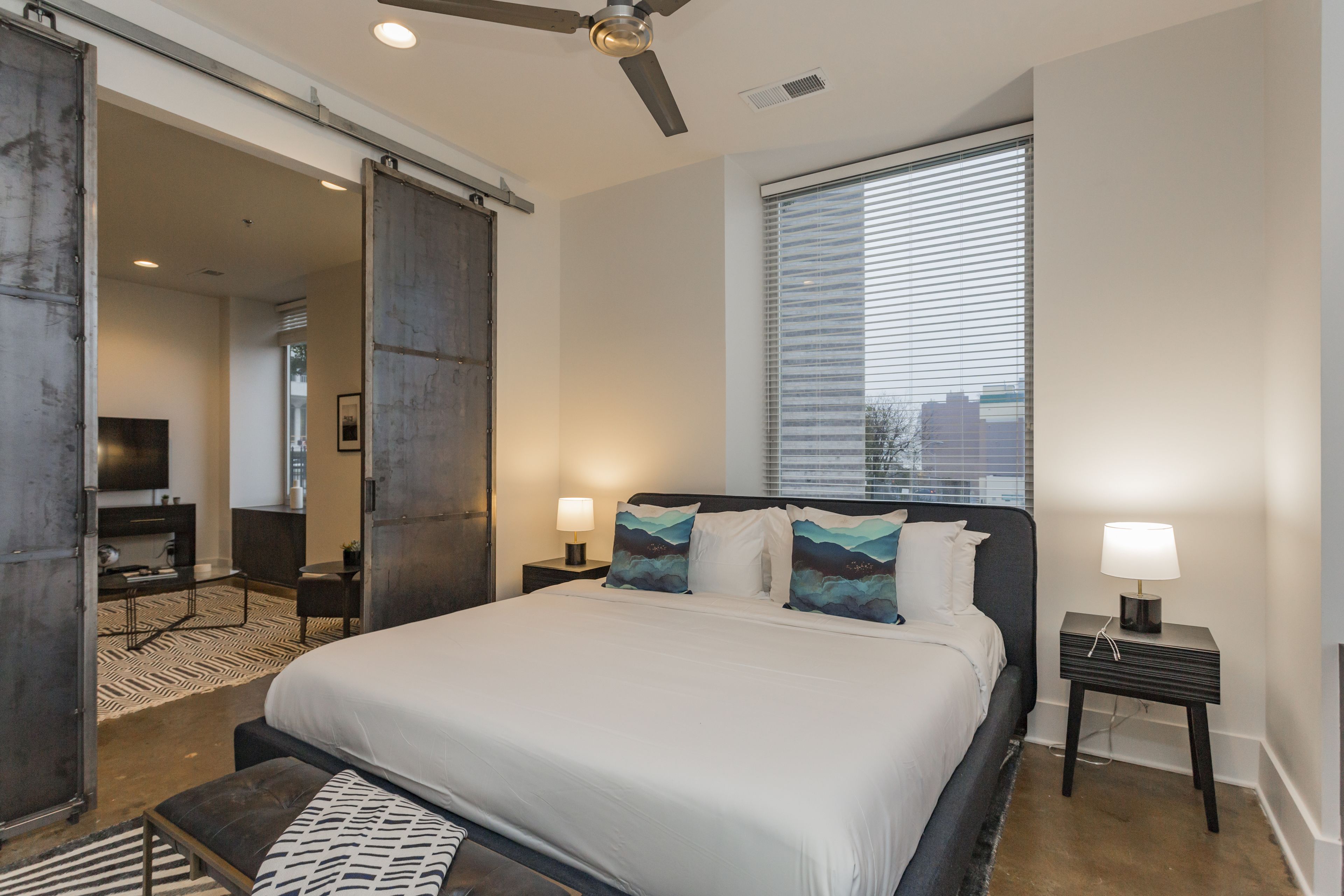 A modern hotel room with a large bed, a ceiling fan, a window with blinds, and a TV in the corner.