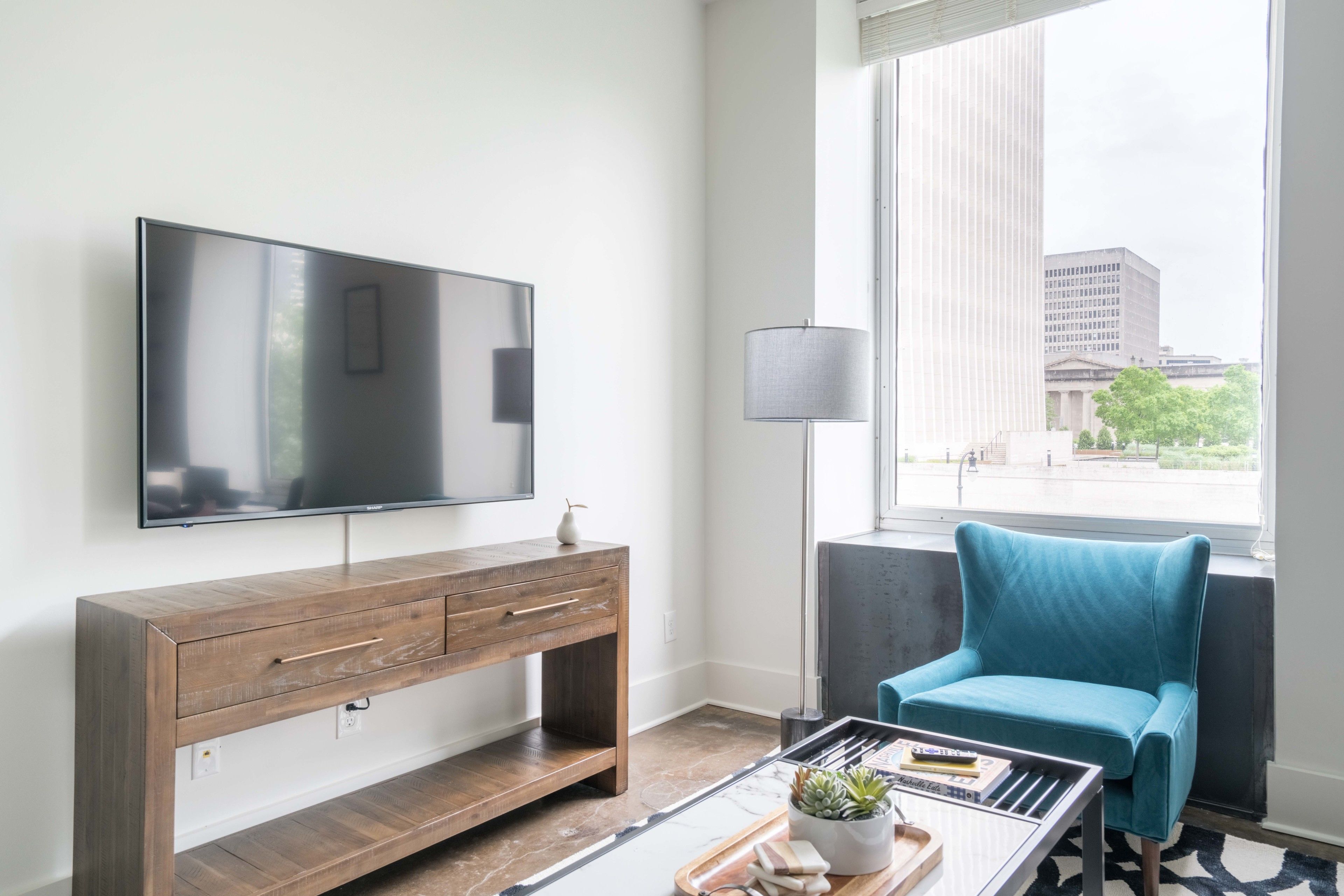 A modern hotel room with a large flat-screen TV, a wooden console table, a teal armchair, and a large window offering a city view.