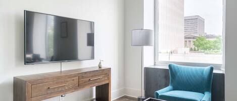 A modern hotel room with a large flat-screen TV, a wooden console table, a teal armchair, and a large window offering a city view.