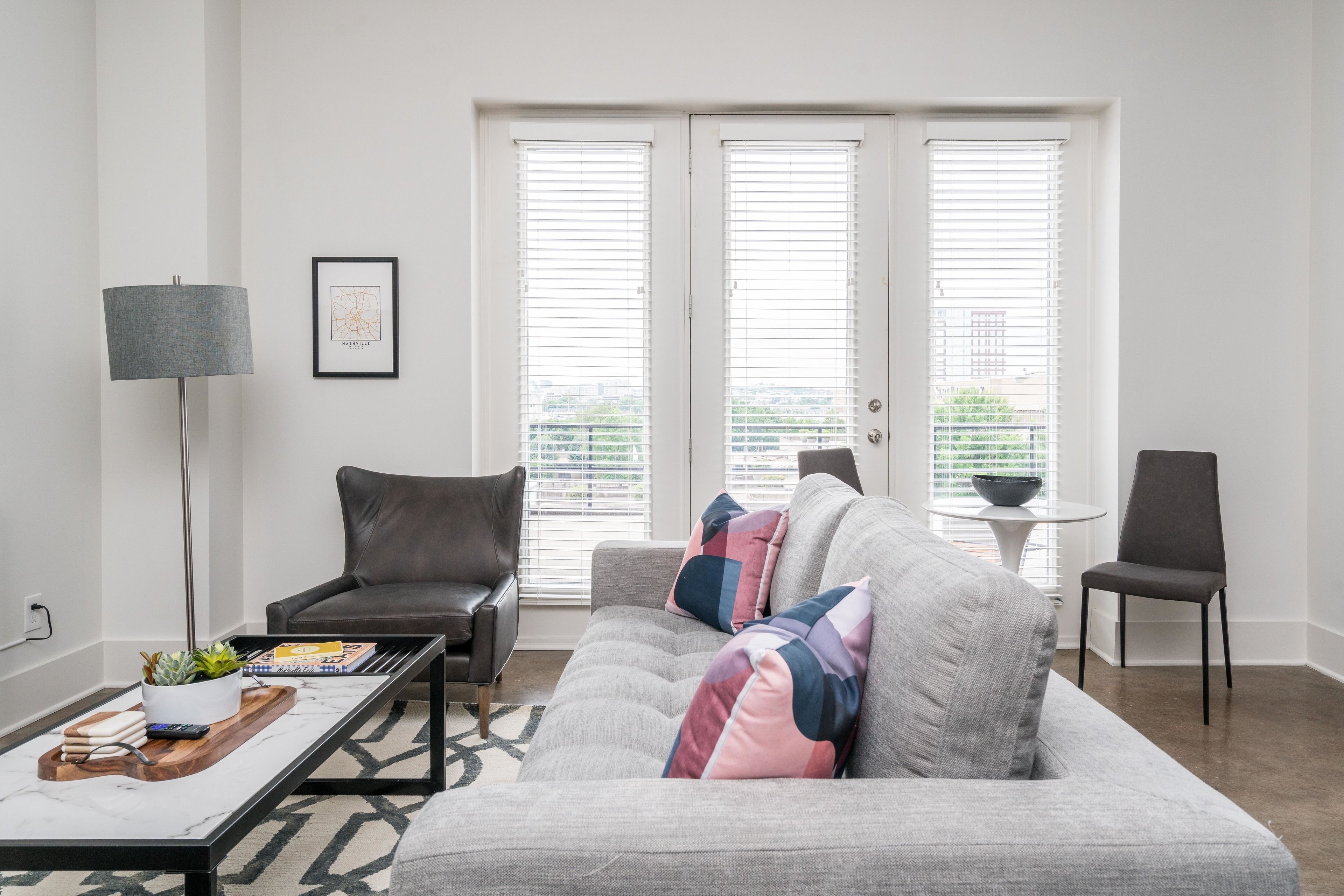 Una sala de estar moderna con un sofá gris, una mesa de centro, una lámpara de pie y una silla. Hay dos ventanas con persianas, un cuadro en la pared y una mesita con un cuenco.