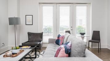 Una sala de estar moderna con un sofá gris, una mesa de centro, una lámpara de pie y una silla. Hay dos ventanas con persianas, un cuadro en la pared y una mesita con un cuenco.