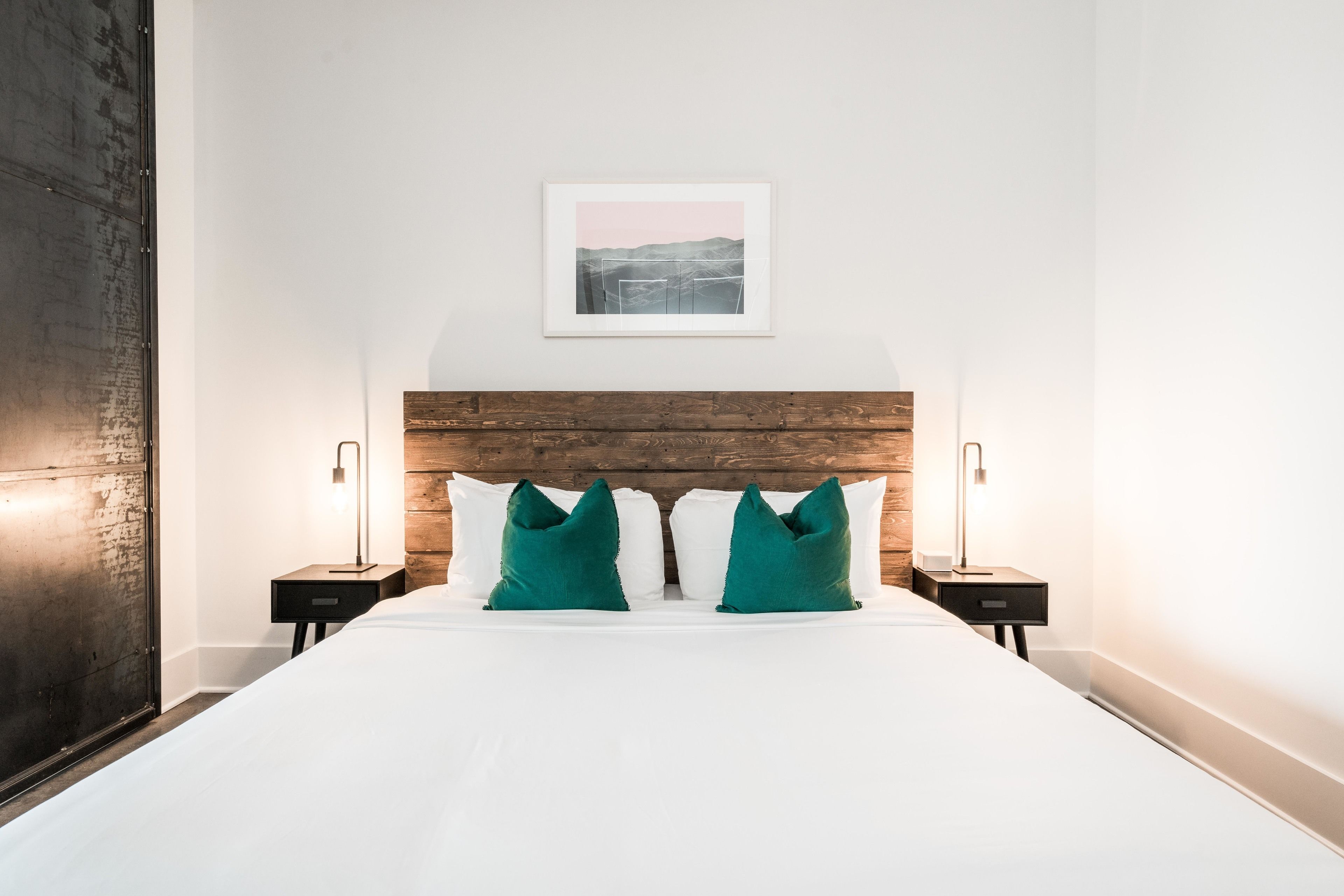 A neatly made bed with white linens and two green pillows, flanked by two bedside tables with lamps, in a room with a wooden headboard and a framed picture above the bed.