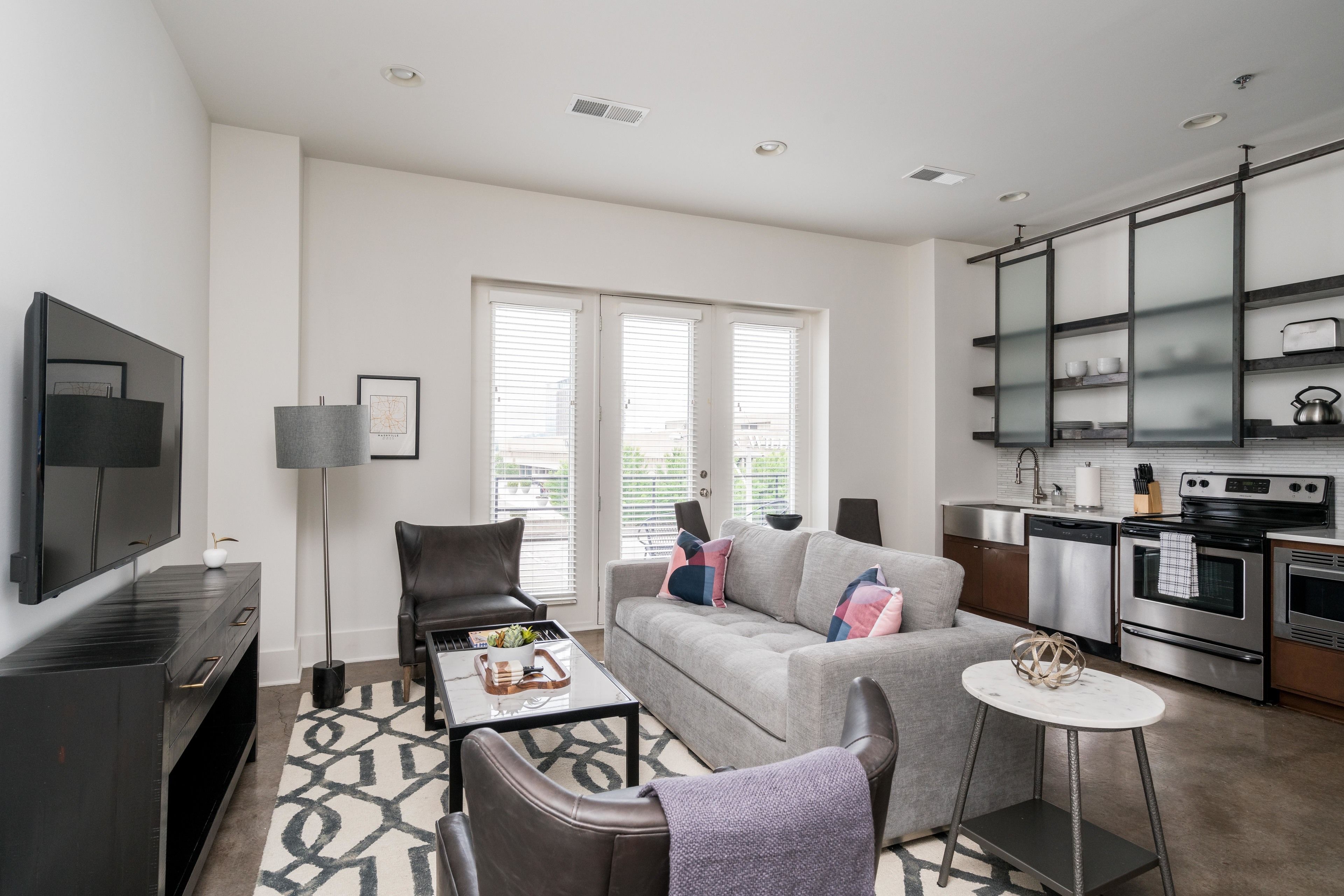 A modern living room with a grey sofa, black leather chairs, a coffee table, and a kitchen area with stainless steel appliances.