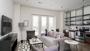 A modern living room with a grey sofa, black leather chairs, a coffee table, and a kitchen area with stainless steel appliances.