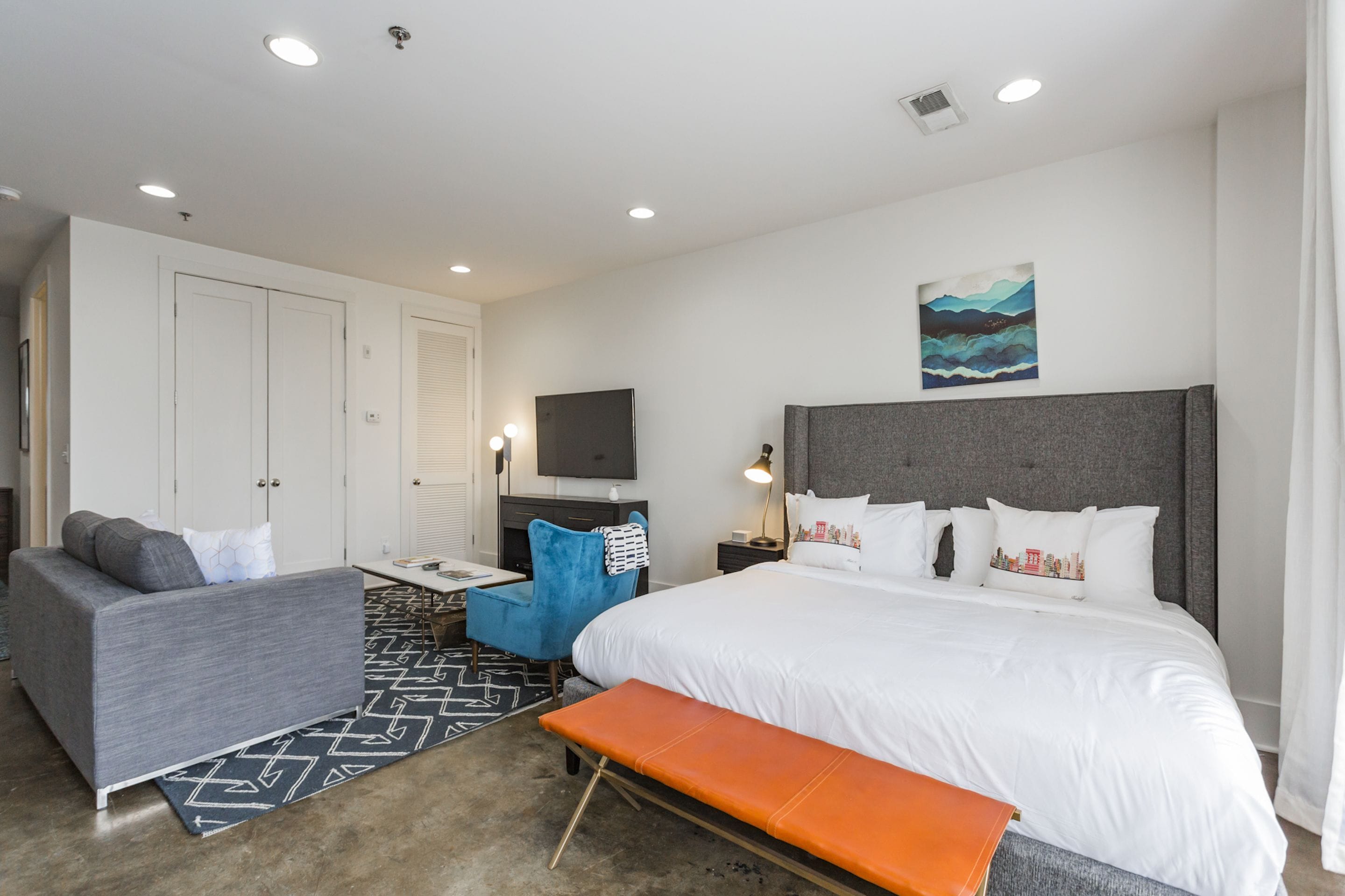 A modern hotel room with a large bed, a grey sofa, a blue armchair, a small desk with a TV, and a painting on the wall.