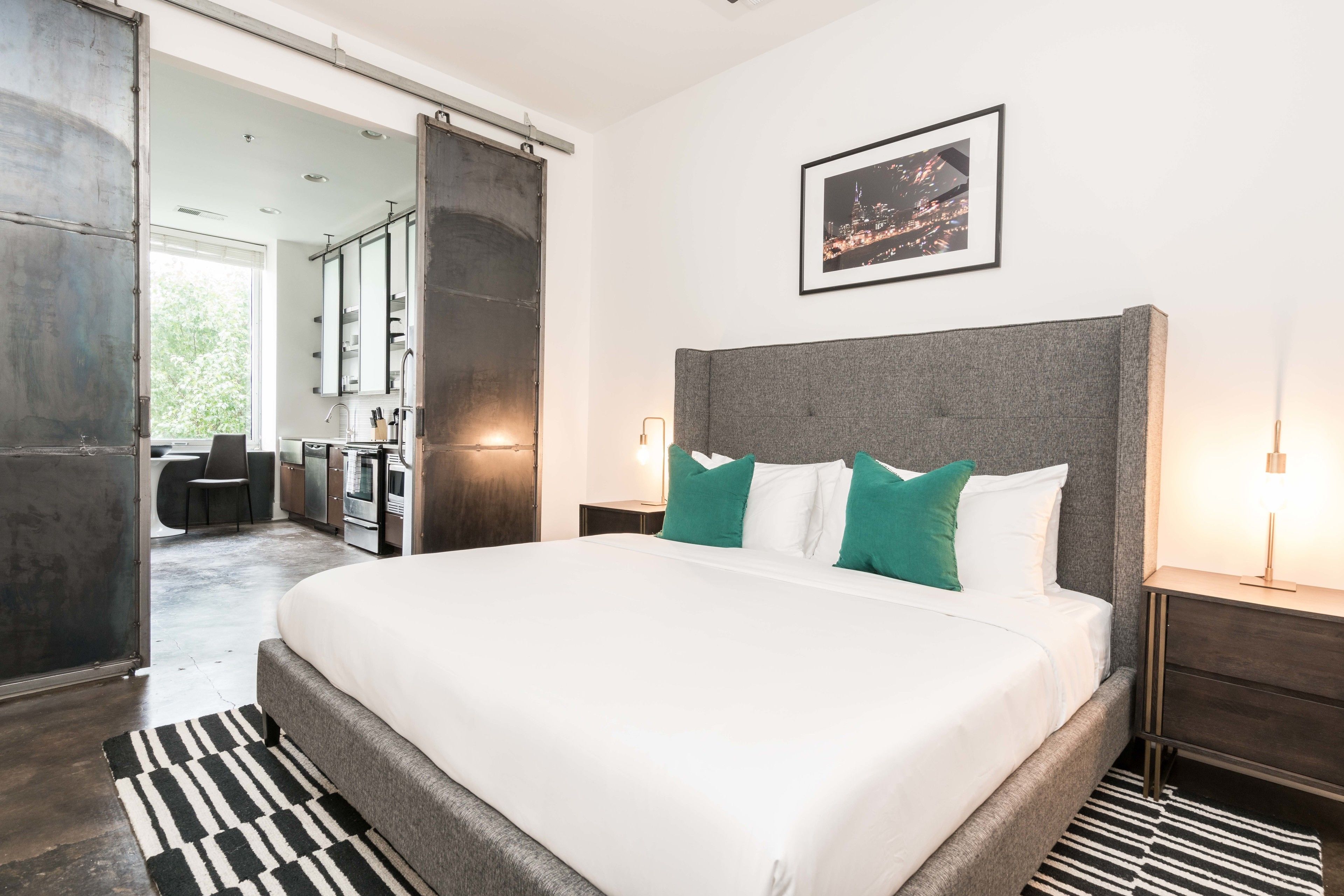 Habitación de hotel moderna con una cama grande, vistas a la ciudad y una puerta corredera que da a un comedor.